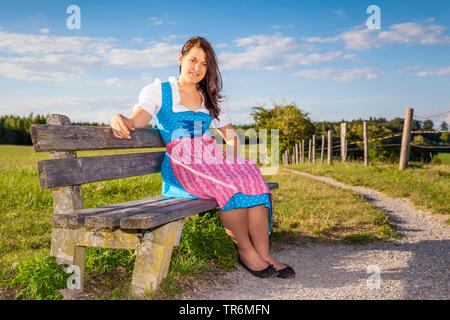 Junge Frau im Dirndl auf einer Bank sitzen auf einer Bergwiese, Deutschland, Bayern Stockfoto