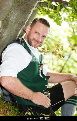 Traditionell gekleidet Bayerische Mann in einem Wald sitzt an einem Baum, Deutschland, Bayern Stockfoto