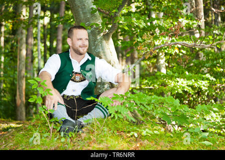 Traditionell gekleidet Bayerische Mann in einem Wald sitzt an einem Baum, Deutschland, Bayern Stockfoto