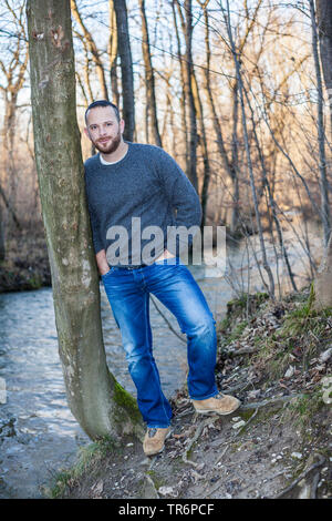 Schöner Mann in der Natur an einem Baumstamm an einem Bach, Deutschland Stockfoto