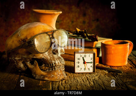 Stilleben mit Schädel mit Sonnenbrille und Wecker, vasevase, Glas, Keramik, Stift, altes Buch, chemische Rosen auf Holz Stockfoto