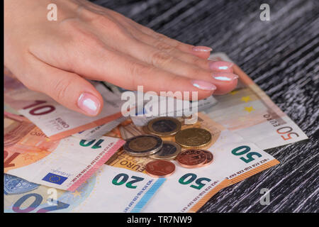 Woman's Hand mit rosa Nägel sind ein Haufen von Euro-Banknoten und -Münzen auf einem schwarzen alten Tisch. Das Konzept der jährlichen Haushaltsplan und zuverlässige Bank Einlagen Stockfoto