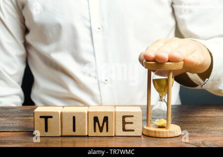 Ein Mann legt Sanduhr in der Nähe von Holz- Bausteine mit dem Wort Zeit. Konzept der Zeit Verwaltung und ordnungsgemäße Verteilung. Planung Wirtschaft und Arbeit. Business y Stockfoto