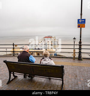 Cromer, Norfolk, Großbritannien. 19. Mai 2019. Ein rentnerehepaar setzte sich auf eine Holzbank vor der Promenade Blick aus Meer und die Pier in der Ansicht auf einem Nebelhaften m Stockfoto