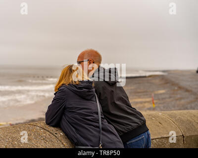 Cromer, Norfolk, Großbritannien. 19. Mai 2019. Ein glückliches Paar mittleren Alters lehnte sich gegen die Wand auf der Promenade beobachten einige Knaben mit einem Surfen Stockfoto