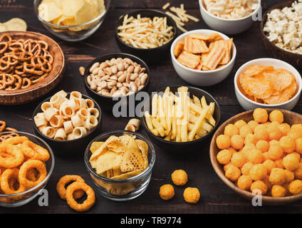 Alle klassischen Kartoffel Snacks mit Erdnüssen, Popcorn und Zwiebelringen und gesalzene Brezeln in Schüssel Platten auf Schwarz. Twirls mit Stöcken und Kartoffelchips Stockfoto