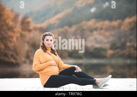 Lächelnd schwangere Frau in Form gestrickter Pullover über Herbst Natur Hintergrund posiert. Mit Blick auf die Kamera. Die Mutterschaft. Mutterschaft. 20. Stockfoto