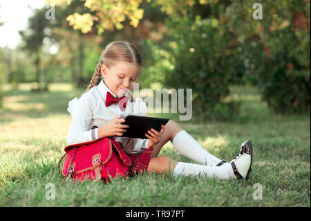 Lächelnd kind Mädchen 6-7 Jahre alten Spielen Tablet im Freien. Tragen Schuluniform. Stockfoto