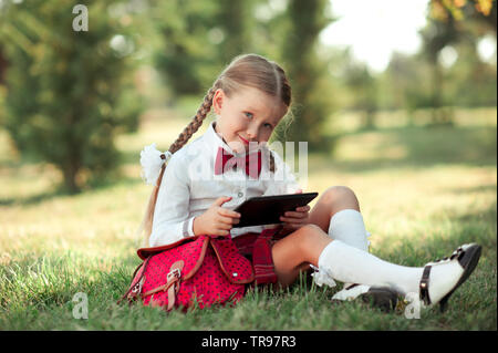 Lustig kind Mädchen 6-8 Jahre alten Holding Tablet im Freien. Mit Blick auf die Kamera. Sitzen in der Wiese. Tragen Schuluniform. Kindheit. Stockfoto