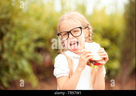 Lustig kind Mädchen essen Sandwich im Freien. Spaß haben. Mit Blick auf die Kamera. Posing über Natur Hintergrund. Gesundes Essen. Kindheit. Stockfoto