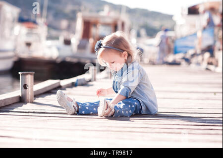 Cute Baby Mädchen 3-4 Jahre alten tragen stilvolle denim Kleidung sitzen auf Holz- Meer Pier im Freien. Verspielt. Kindheit. Sommer Saison. Stockfoto