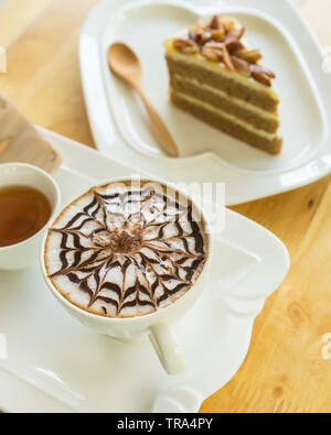 Heißer Kaffee spät in weißem Glas auf Holztisch, verschwommenes Kuchen und Löffel aus Holz, Stockfoto