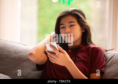 Biracial jugendlich Mädchen sitzen auf grau Couch durch große Panoramafenster, auf Smartphone suchen Stockfoto