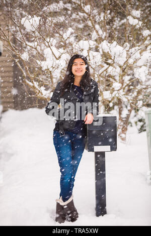 Biracial jugendlich Mädchen in Blue Jeans und schwarze Lederjacke lehnte sich gegen Mailbox im Winter Schneesturm Stockfoto