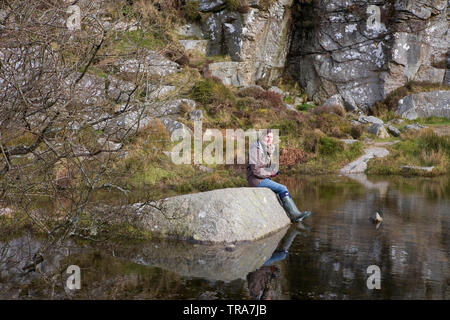 Junge Frau sitzt auf einem Felsen am Pool im Haytor Steinbrüche, Haytor, Dartmoor, Devon, England. MODEL RELEASED Stockfoto