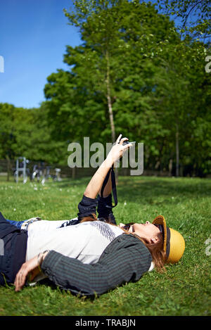 Zwei weibliche beste Freunde mit in das Gras zusammen im Park im Sommer Sonnenschein einen selfie mit einer Tasche für Kamera Stockfoto