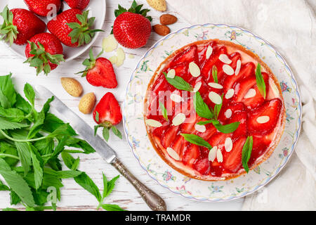 Hausgemachten leckeren Erdbeerkuchen mit Sahne, Pudding oder mit Minze und Mandeln Blütenblätter in eine Platte auf eine weiße Holztisch. Dessert für Stockfoto