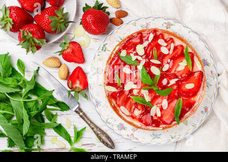 Hausgemachten leckeren Erdbeerkuchen mit Sahne, Pudding oder mit Minze und Mandeln Blütenblätter in eine Platte auf eine weiße Holztisch. Dessert für Stockfoto