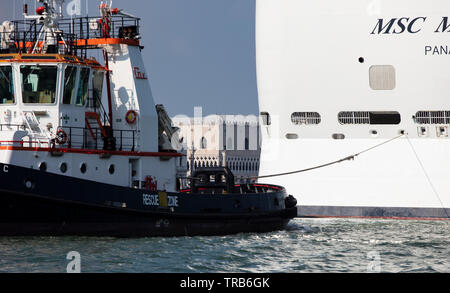 Ein Kreuzfahrtschiff in den Giudecca Kanal mit einem Tauziehen, klein erscheinen die Gebäude entlang der historischen Waterfront von Venedig wie es geht. Stockfoto