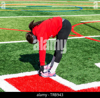 Ein junges Mädchen Leichtathletik runner ist Stretching ihr Kniesehnen im Winter outdoor Praxis tragen ein Rotes Sweatshirt und schwarze Spandex. Stockfoto