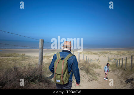 Ein Mann laufen über eine Sanddüne im hellen Sonnenschein trägt blaue Jeans und einen Rucksack und einen Strohhut Stockfoto