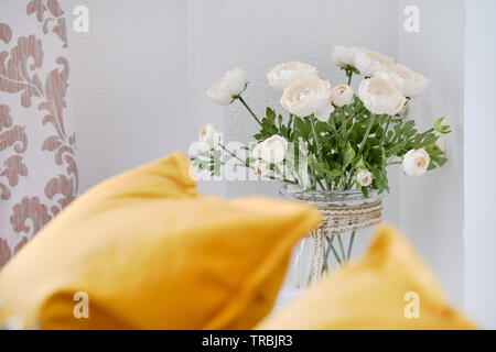 Gelbe Kissen auf der Couch im Wohnzimmer, am wunderschönen Blumen auf einem dünnen Stamm in Glasvase weißen Ranunkeln in der Nähe der Wall niedlich Dekoration Zuhause Stockfoto