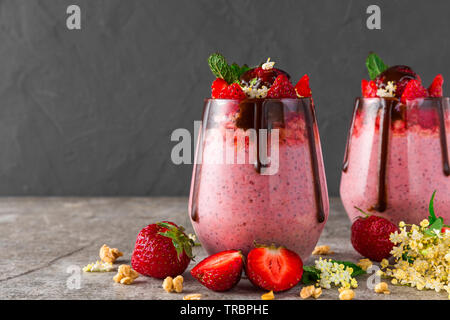 Zwei Gläser Erdbeer Chia Samen Pudding mit Schokolade, frische Beeren, Müsli, elderflowers und Minze für gesundes leckeres Frühstück auf Beton zurück Stockfoto