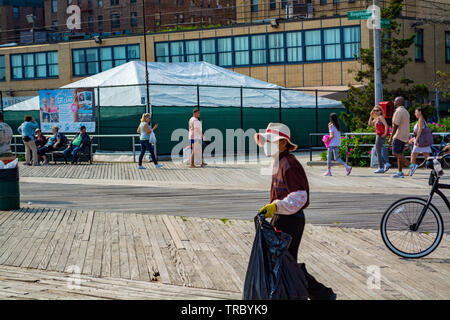 Asiatischer Mann mit Maske ist das Sammeln von leeren Plastikflaschen auf der Strandpromenade, am frühen Nachmittag. Stockfoto