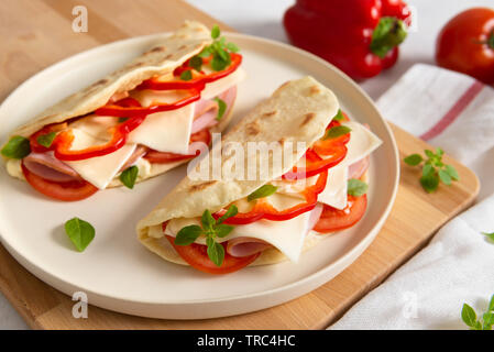 Italienisches Piadina Romagnola Fladenbrot mit Paprika, Tomaten, Schinken, Käse und Basilikum auf der Platte auf weißem Holz- Hintergrund. Close Up. Ita Stockfoto