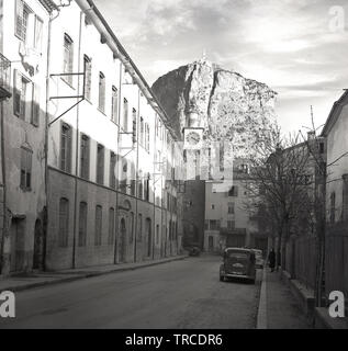 1950s, historisch, Blick auf eine ruhige, verlassene Straße in der Stadt Sisteron, Frankreich, mit einem Auto der Zeit. Die Stadt ist bekannt als das "Tor zur Provence", da sie zwischen zwei langen Bergketten liegt, von denen eine auf dem Bild zu sehen ist und über der Stadt aufragt. Stockfoto