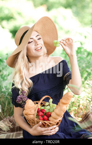 Lächelnde Mädchen 24-29 Jahre alten mit Picknick im Park. Holding Weidenkorb mit Früchten und Brot. Tragen Strohhut und elegantes blaues Kleid im Freien. Summe Stockfoto