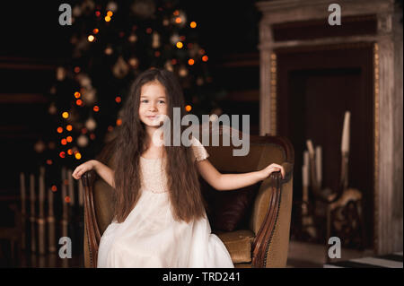 Lächelnden Mädchen 4-5 Jahre alten tragen Trendfarbe weiß Kleid in vintage Stuhl im Zimmer über Weihnachten Baum mit Lichtern. Mit Blick auf die Kamera. Urlaub Stockfoto