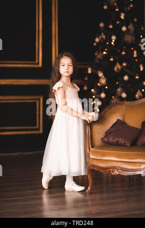 Cute Baby Mädchen 4-5 Jahre alten tragen elegantes weißes Kleid über Weihnachten Lichter im Raum darstellen. Mit Blick auf die Kamera. Urlaubszeit. Stockfoto