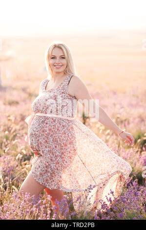 Lächelnd schwangere Frau tragen elegantes Kleid mit Blumenmuster in der Wiese. Mit Blick auf die Kamera. Die Mutterschaft. Mutterschaft. Stockfoto