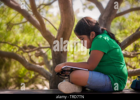 Nette junge Mädchen sitzen im Schatten eines Baumes, auf Ihr Buch lesen und lassen ihrer Fantasie freien unter der warmen Sonne Kaliforniens laufen konzentriert. Stockfoto