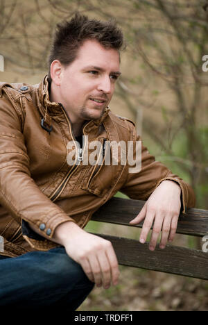 Portrait einer jungen sympathischen Mann in einer Lederjacke auf einer Parkbank gekleidet Stockfoto