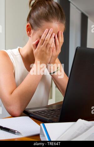 Portrait einer jungen Frau verzweifelt an einem Schreibtisch, die ihr Gesicht mit den Händen Stockfoto