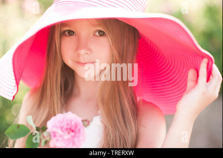 Lächelnde blonde Kind Mädchen 4-5 Jahre alten tragen große rosa Hut Holding stieg im Freien. Mit Blick auf die Kamera. Kindheit. Sommer. Stockfoto