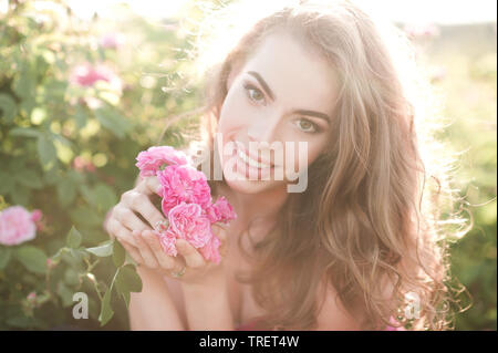 Lächelnd schöne blonde Frau 24-29 Jahre alten Holding Rose Blume in der Wiese im Freien posieren. Mit Blick auf die Kamera. Sommer. Stockfoto