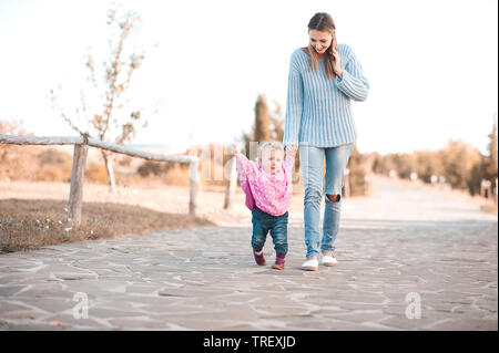 Baby girl 1 Jahr alt, erste Schritte halten Mütter hand Walking im Park. Legere Kleidung Herbst im Freien. Die Mutterschaft. Stockfoto