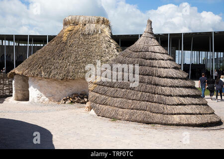 Von außen neu Steinzeit Steinzeit Hütte/stoneage Hütten mit Reetdach/Dächer während Touristen herum gehen. Dorf Ausstellung; Visitor Center Stonehenge/Stone Henge. Amesbury, Wiltshire, UK (109) Stockfoto