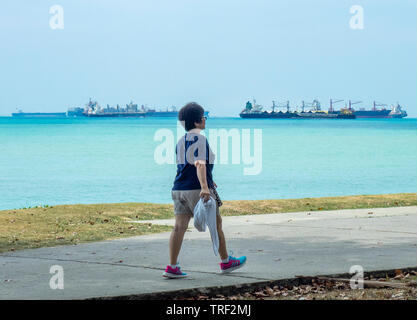 Frau mittleren Alters entlang einem Pfad und Fracht Containerschiffe im Hintergrund East Coast Park Singapur. Stockfoto