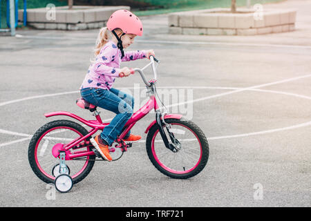 Portrait von lächelnden Kaukasischen Vorschüler Mädchen, rosa Fahrrad in Helm auf Hinterhof Straße draußen auf Frühling Sommer Tag. Saisonale kind Aktivitäten Stockfoto