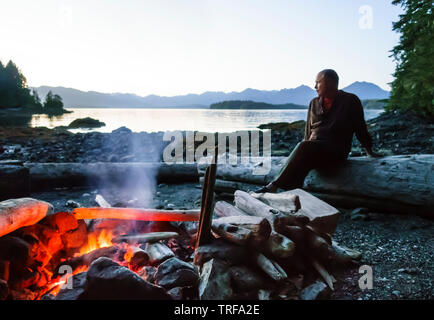 Mann sitzt auf Strand am Lagerfeuer in der Wildnis, Broken Group Islands, Barkley Sound, Pacific Rim National Park, Kanada. Selektiver Fokus auf Feuer. Stockfoto