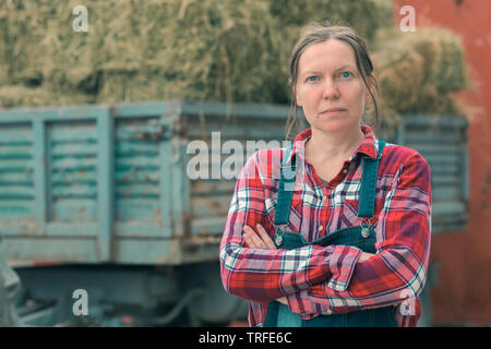 Frau Bauer vor ladewagen posieren. Portrait von Frau Landarbeiter in Plaid Shirt und bib Overalls vom Traktor Anhänger gefüllt mit Molkerei weit Stockfoto