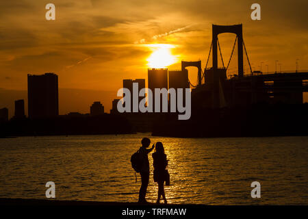 Ein junges Paar genießt den Blick auf die Rainbow Bridge von einem Strand in Odaiba, Tokio, Japan. Stockfoto