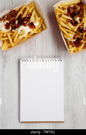 Fast food: Pommes Frites mit Käsesoße und Speck in Papier, Kartons, leere Notepad auf weißem Holz- Hintergrund, Ansicht von oben. Stockfoto