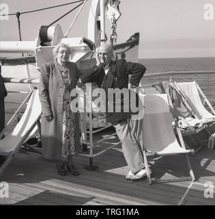 1950, historische, ein älteres Paar auf einer Kreuzfahrt stehend auf dem Deck der P&O Ocean Liner, SS Chusan. Die Frau trägt ein langes Kleid, mit ein Mantel, während der Mann mehr passend für Kreuzfahrt in einem Blazer und deck Schuhe gekleidet ist und das Rauchen einer Pfeife. Stockfoto