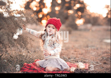 Cute kid Mädchen 4-5 Jahre alten schmücken Weihnachtsbaum im Freien. Tragen Strickmütze, Pullover und Rock. Mit Blick auf die Kamera. Stockfoto