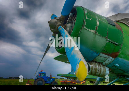 Bunte blau und gelb Propeller auf einer Vintage grüne Ebene geparkt, im Freien in einem ländlichen Gebiet mit einem Traktor im Hintergrund sichtbar in der Nähe zu de Stockfoto